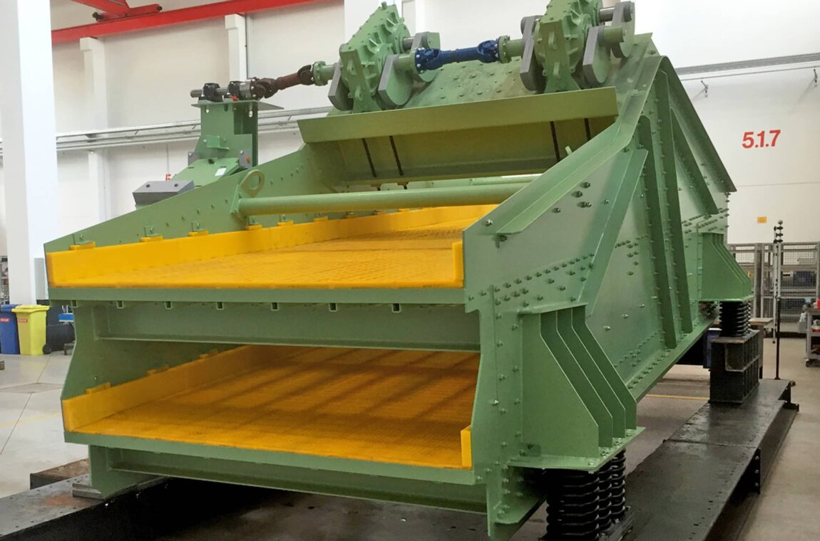 Linear vibratory screening machine HG 22-5_76-II WS - discharge side. The machine is located in the production hall and can be seen as a whole. It is a machine with 2 screen decks and yellow polyurethane screen linings.